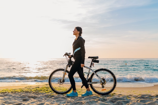Jeune femme mince attrayante à vélo, sports sur la plage du lever du soleil du matin dans des vêtements de sport de remise en forme de sport, mode de vie sain et actif, souriant heureux de s'amuser