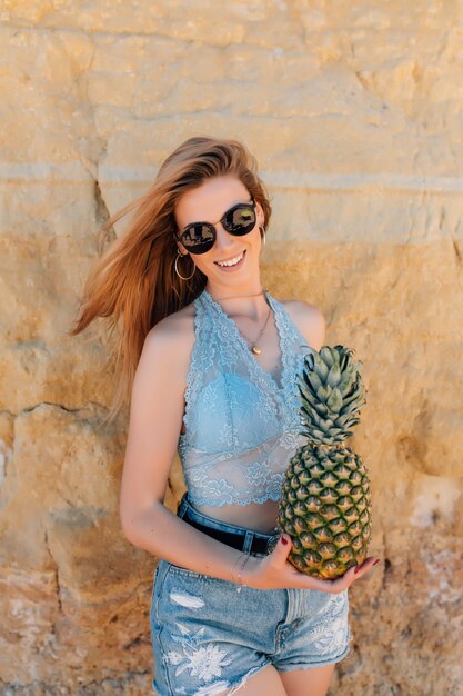 Jeune femme mince en bikini et lunettes de soleil tenant ananas frais près de la mer
