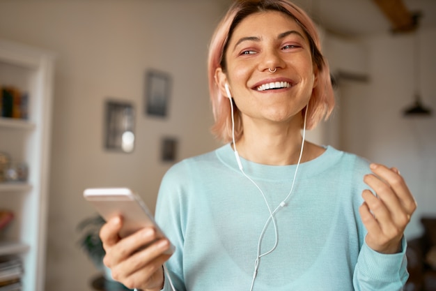 Jeune femme à la mode aux cheveux roses, profitant de la communication en ligne, parlant à un ami en vidéoconférence à l'aide d'écouteurs et wifi sur téléphone intelligent.