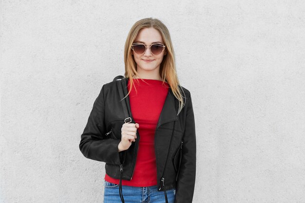 Jeune femme à la mode dans de grandes lunettes de soleil à la mode, pull rouge, jeans et manteau en cuir tenant un sac à dos pour les cours à l'université