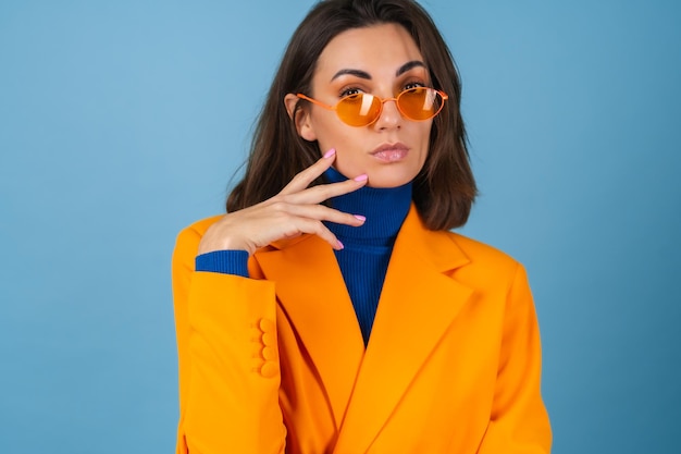 Jeune femme à la mode dans une veste surdimensionnée et au genou sur un mur bleu dans des lunettes orange vif élégantes posant