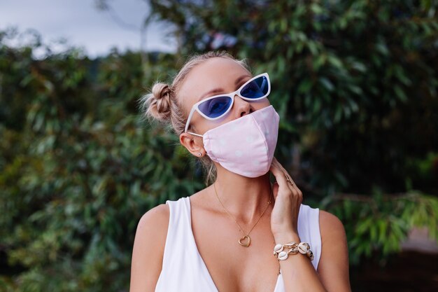 Jeune femme de mode en haut court blanc, lunettes de soleil et masque médical blanc dans la nature