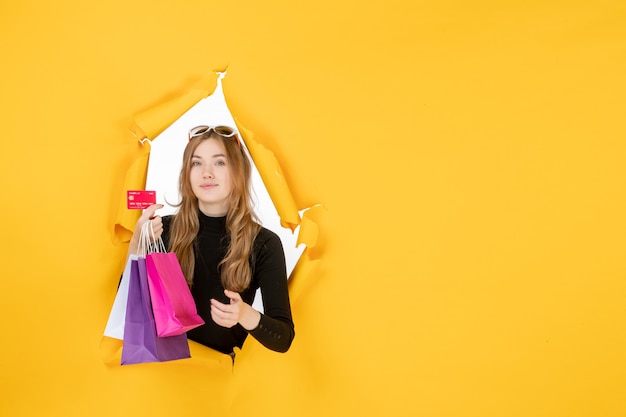 Photo gratuite jeune femme de mode tenant des sacs à provisions et une carte de crédit à travers un trou de papier déchiré dans le mur