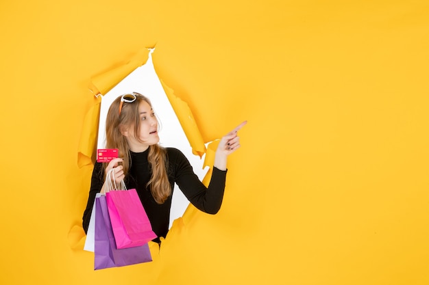 Jeune femme de mode tenant des sacs à provisions et une carte de crédit à travers un trou de papier déchiré dans le mur