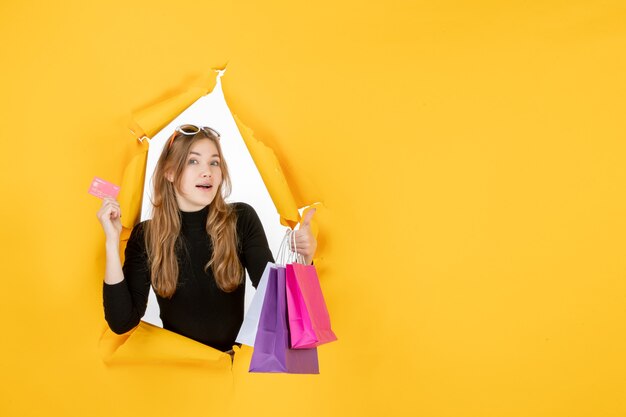 Jeune femme de mode tenant des sacs à provisions et une carte de crédit à travers un trou de papier déchiré dans le mur