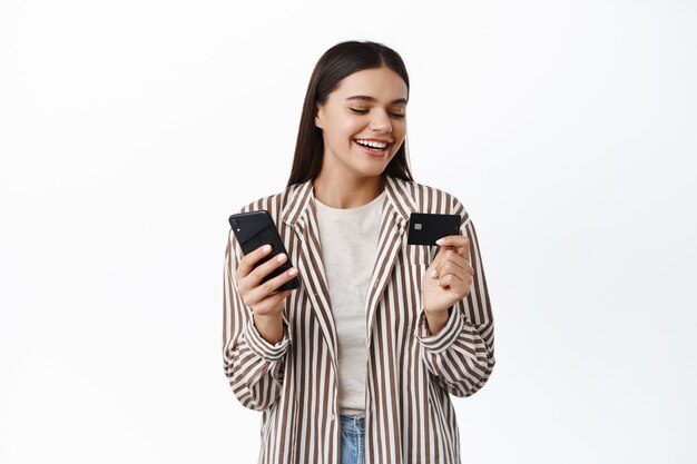 Jeune femme moderne élégante souriante, regardant une carte de crédit en plastique, payant en ligne avec un téléphone portable, faisant des achats dans une application pour smartphone, mur blanc