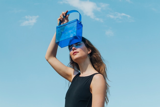 Jeune femme moderne protégeant ses yeux à travers un sac en plastique bleu contre le ciel