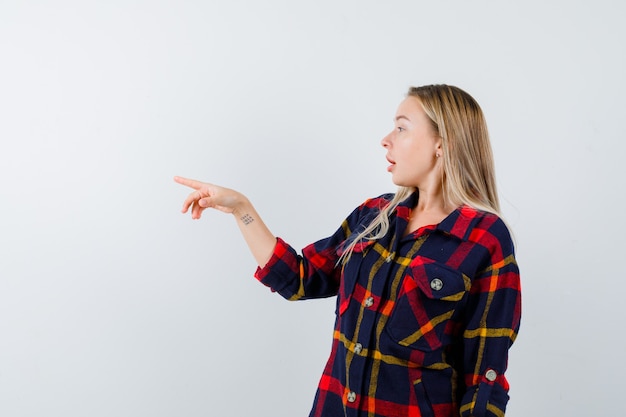 Jeune femme montrant le côté gauche en chemise à carreaux et l'air étonné. vue de face.