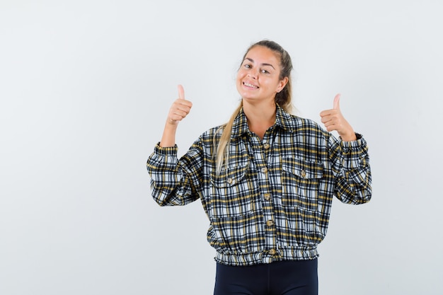 Jeune femme montrant deux pouces vers le haut en chemise à carreaux et à la vue de face, joyeuse.