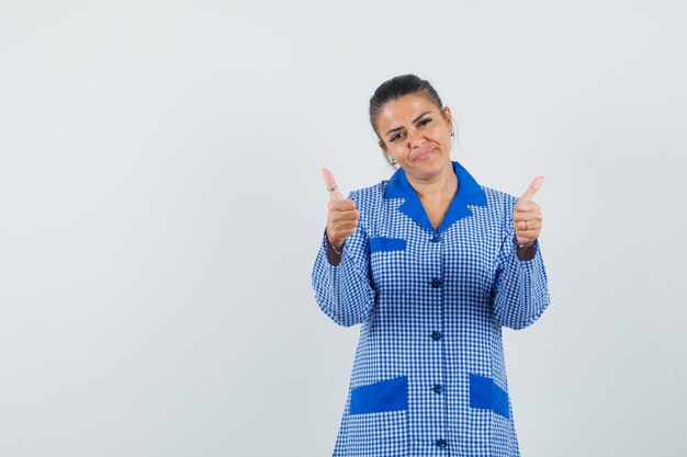 Jeune femme montrant deux pouces vers le haut en chemise de pyjama vichy bleu et à la jolie. vue de face.