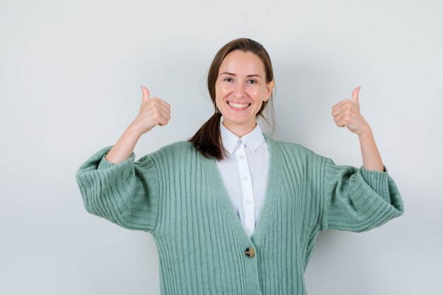 Jeune femme montrant un double coup de pouce en chemisier, cardigan et l'air joyeux. vue de face.