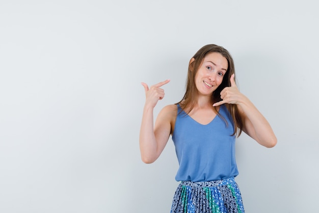 Jeune femme montrant le geste d'appel téléphonique tout en pointant sur elle-même en chemisier bleu, jupe et à la vue de face, confiant.