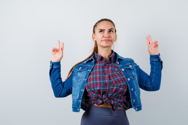 Jeune femme montrant un geste d'arme à feu en chemise à carreaux, veste, pantalon et l'air sérieux. vue de face.