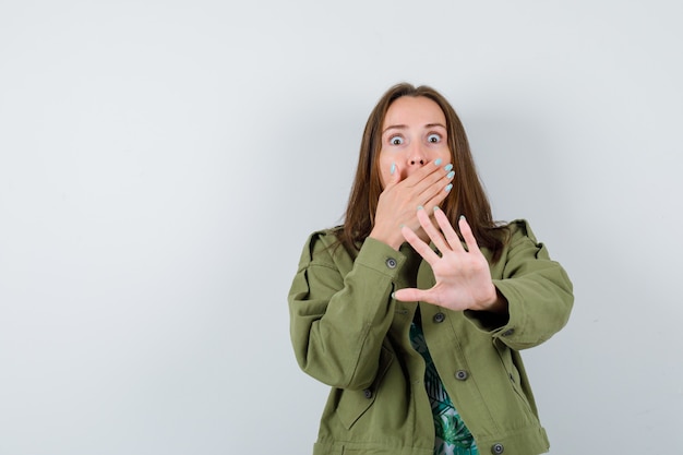 Jeune femme montrant un geste d'arrêt, la main sur la bouche en veste verte et l'air terrifiée, vue de face.