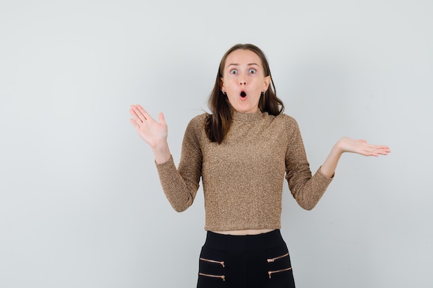 Jeune femme montrant le geste d'arrêt avec la main et tenant une autre main comme présentant quelque chose en pull doré et pantalon noir et à la surprise, vue de face.