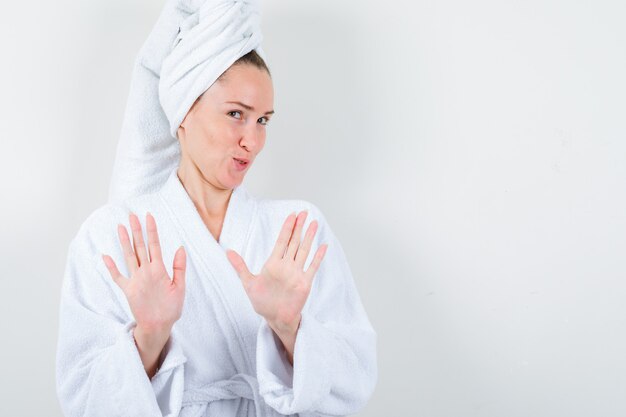 Jeune femme montrant le geste d'arrêt en peignoir blanc, serviette et à la vue sensible, de face.