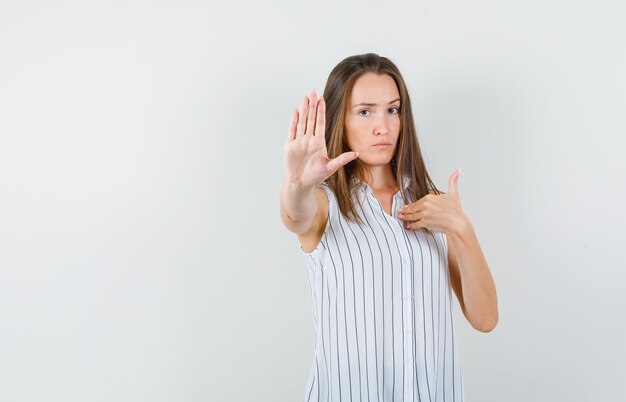 Jeune femme montrant un geste d'arrêt tout en faisant un geste 'moi?' en t-shirt et à la recherche sérieuse, vue de face.