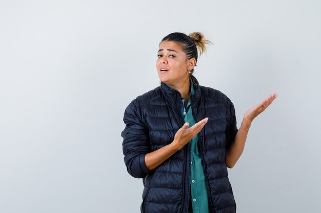 Jeune femme montrant un geste de bienvenue en chemise, doudoune et semblant confuse, vue de face.