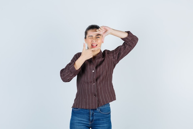 Jeune femme montrant un geste de cadre, un clin d'œil en chemise rayée, un jean et l'air heureux. vue de face.