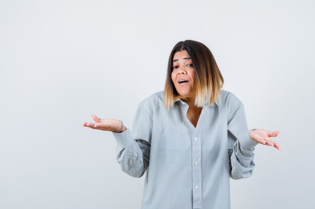 Jeune femme montrant un geste de doute en chemise surdimensionnée et semblant excitée. vue de face.