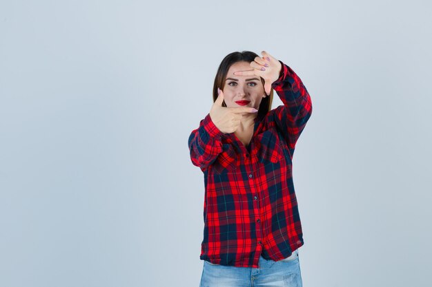 Jeune femme montrant le geste du cadre en chemise à carreaux, jeans et l'air heureux, vue de face.