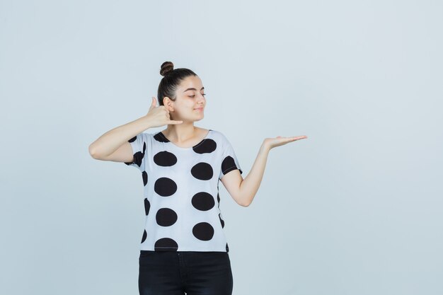 Jeune femme montrant le geste du téléphone tout en faisant semblant de tenir quelque chose en t-shirt, jeans et à la joyeuse vue de face.