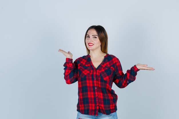 Jeune femme montrant un geste d'écailles en chemise à carreaux et regardant jolly , vue de face.