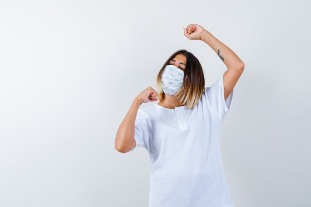 Jeune femme montrant le geste gagnant en t-shirt, masque et à la chance. vue de face.