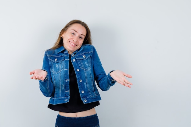 Jeune femme montrant un geste impuissant en blouse, veste et semblant optimiste, vue de face.