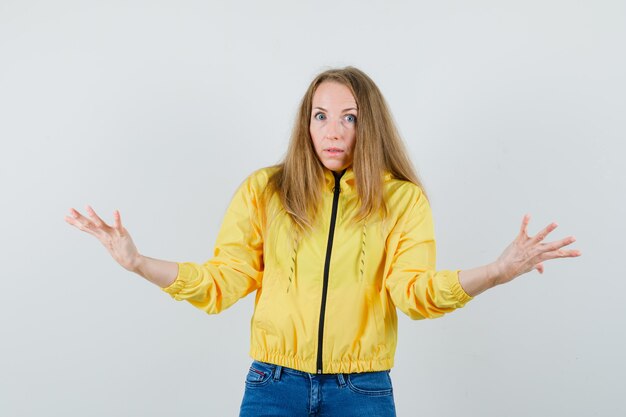 Jeune femme montrant un geste impuissant en blouson aviateur jaune et jean bleu et à la vue douteuse, de face.