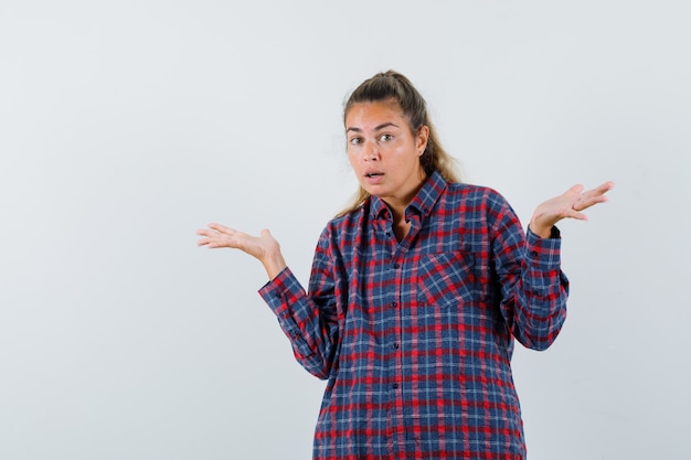 Jeune femme montrant un geste impuissant en chemise à carreaux et à la vue confuse, de face.