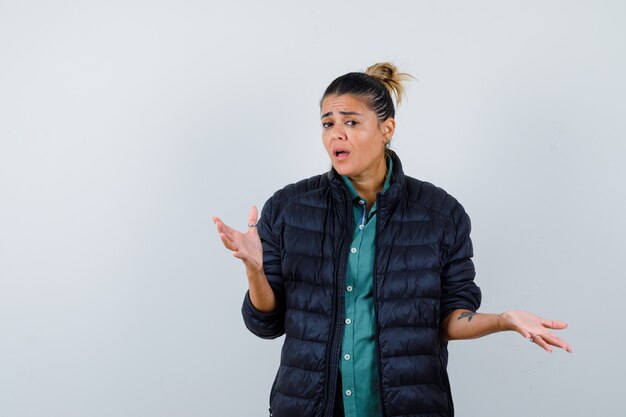 Jeune femme montrant un geste impuissant en chemise, doudoune et à la perplexité. vue de face.