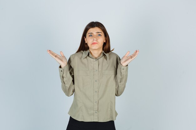 Jeune femme montrant un geste impuissant, courbant les lèvres en chemise, jupe et l'air perplexe, vue de face.