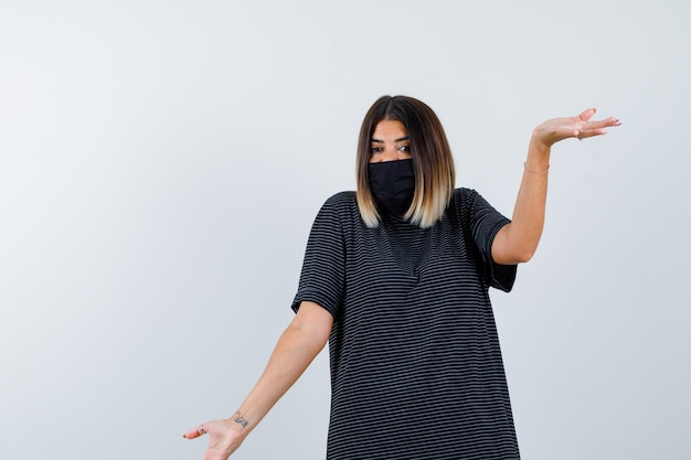 Jeune femme montrant un geste impuissant en robe noire, masque noir et à la perplexité. vue de face.