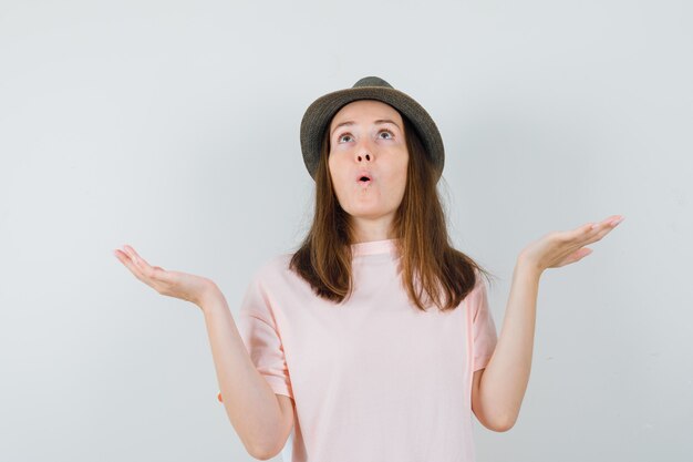 Jeune femme montrant un geste impuissant en t-shirt rose, chapeau et à la perplexité. vue de face.