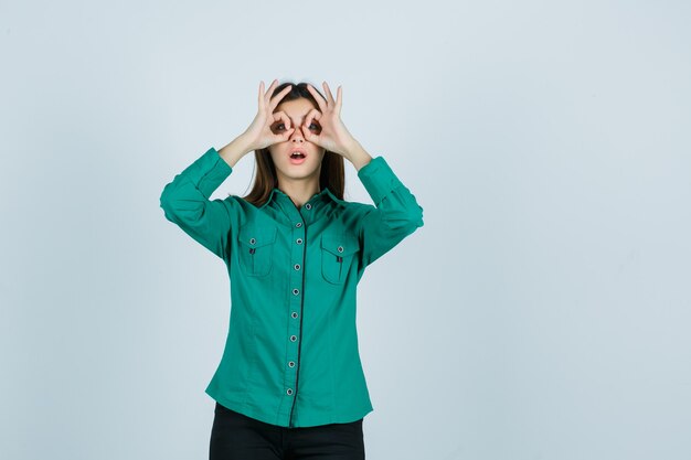 Jeune femme montrant le geste de lunettes en chemise verte et à la recherche de surprise. vue de face.