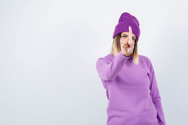 Jeune femme montrant un geste minute en pull violet, bonnet et l'air confiant. vue de face.