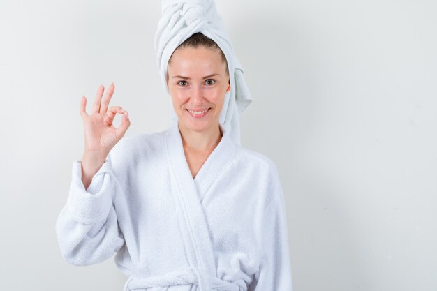 Jeune femme montrant le geste ok en peignoir blanc, serviette et à la vue de face, heureux.