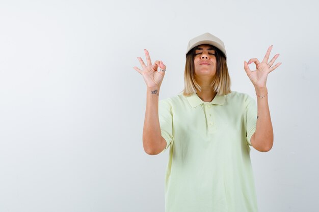 Jeune femme montrant un geste ok sur les yeux en t-shirt, casquette et à la confiance. vue de face.