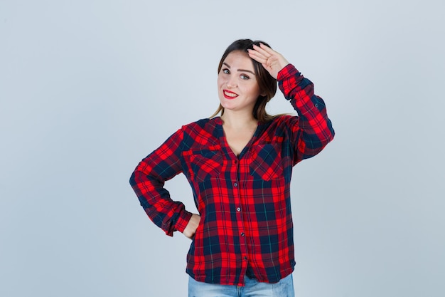 Jeune femme montrant un geste de salut en chemise à carreaux et l'air gaie, vue de face.