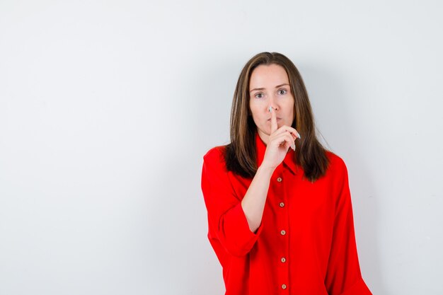 Jeune femme montrant un geste de silence en blouse rouge et semblant sérieuse, vue de face.