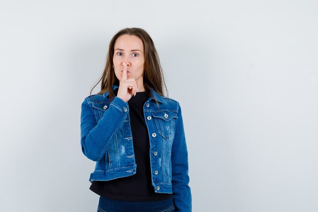 Jeune femme montrant un geste de silence en blouse, veste et l'air confiant. vue de face.