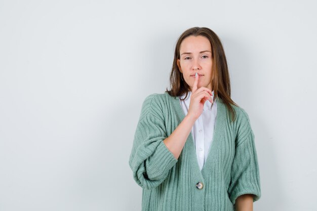 Jeune femme montrant un geste de silence en chemise, cardigan et semblant insatisfaite, vue de face.