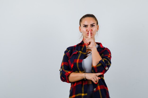 Jeune femme montrant un geste de silence en haut, chemise à carreaux et semblant sérieuse, vue de face.