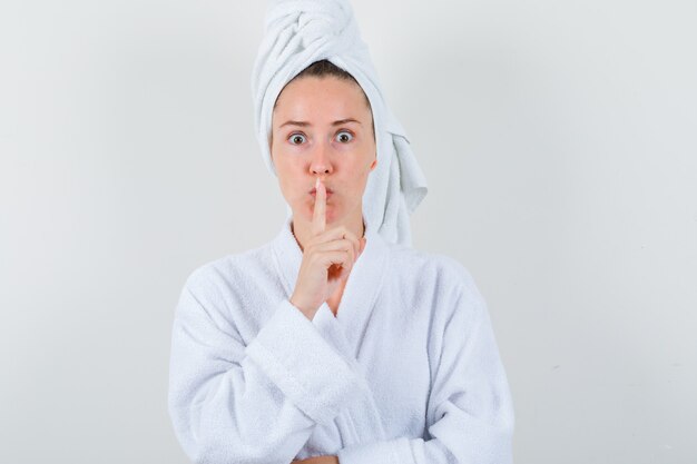 Jeune femme montrant le geste de silence en peignoir blanc, serviette et à la prudence. vue de face.