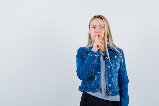 Jeune femme montrant un geste de silence en t-shirt, veste en jean, jupe et l'air confiant