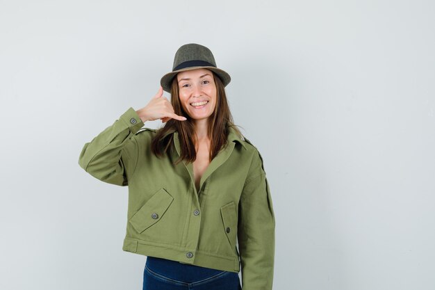 Jeune femme montrant le geste de téléphone dans le chapeau de pantalon de veste et à la joyeuse