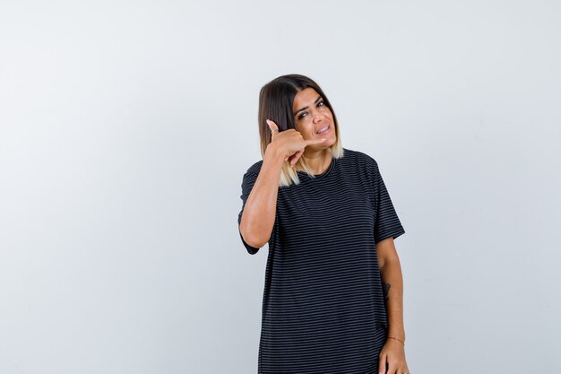 Jeune femme montrant le geste de téléphone en robe de polo et à la vue de face, joyeuse.