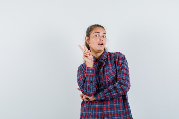 Jeune femme montrant le geste de la victoire en chemise à carreaux et l'air confiant. vue de face.