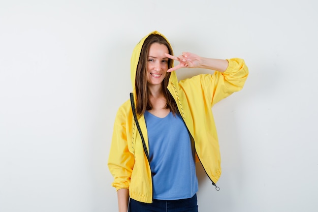 Jeune femme montrant un geste de victoire près de l'œil en t-shirt, veste et l'air confiant, vue de face.